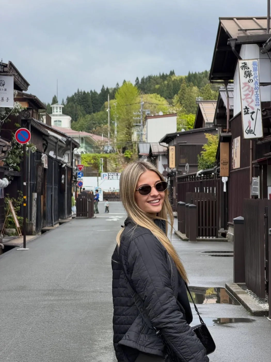 Emi con campera negra y anteojos de sol en una calle con casas de madera