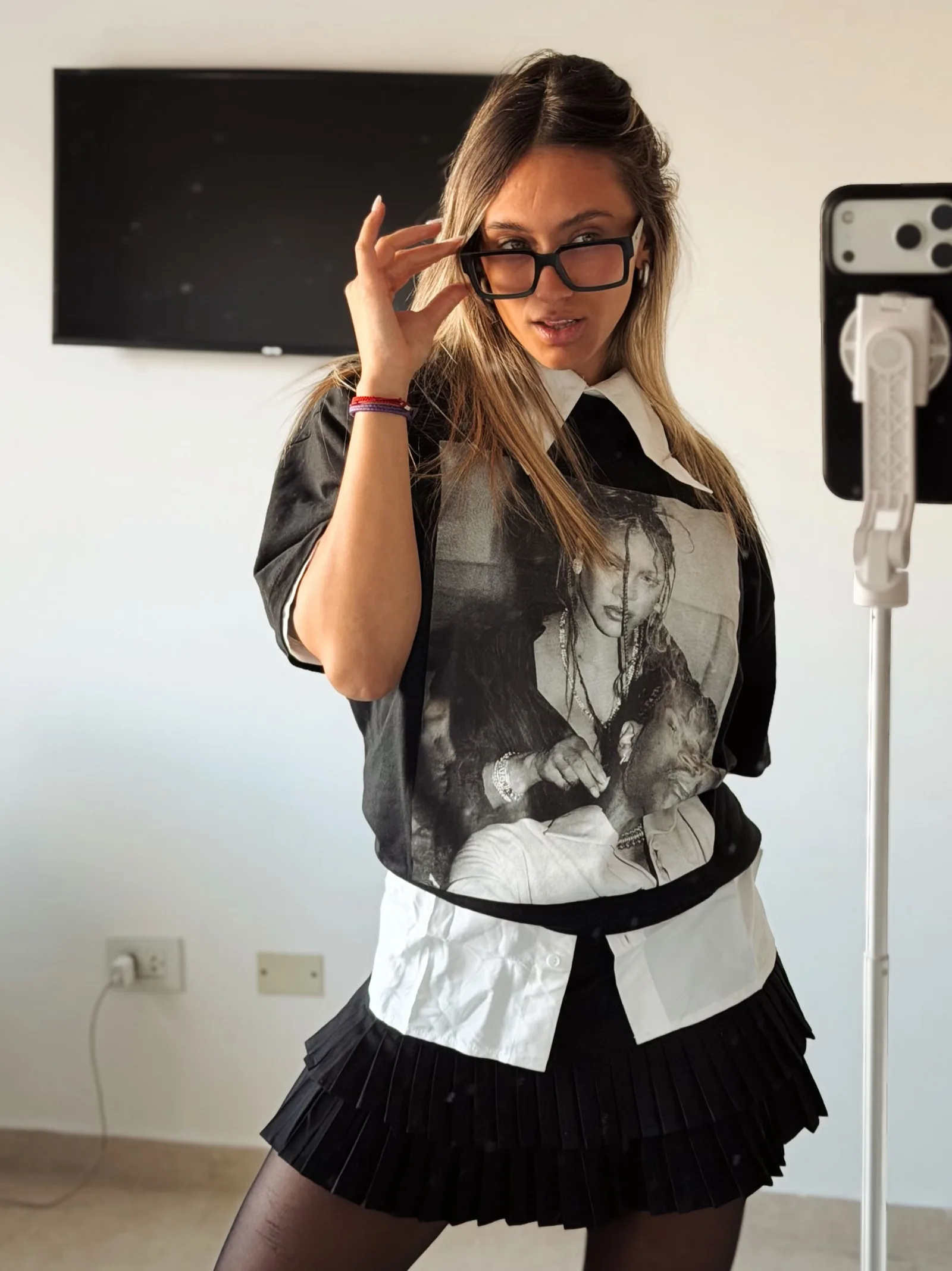 Marti Rinaldi con anteojos, remera estampada, camisa blanca y pollera tableada negra
